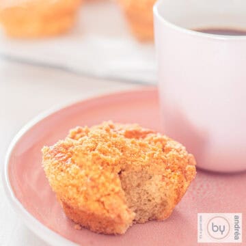 A square photo of a pink plate with a cup of coffee and a speculaas muffin showing the inside. A cup of coffee is in the background.