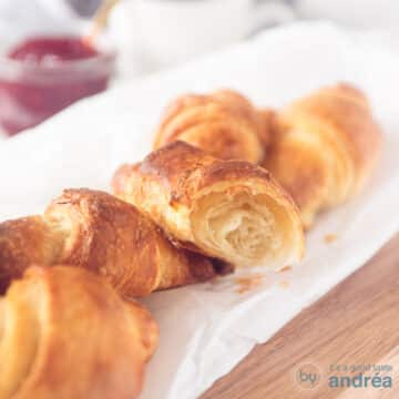 A square photo with a baking paper on a wooden board with four croissants and a half croissant where you can clearly see the fluffy inside.