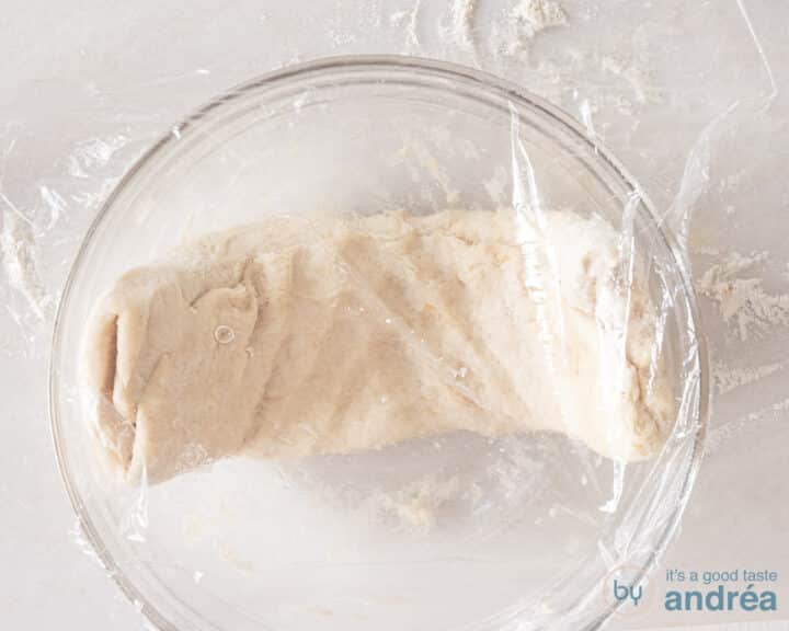 A thick piece of dough is rising in a glass bowl covered with plastic foil.