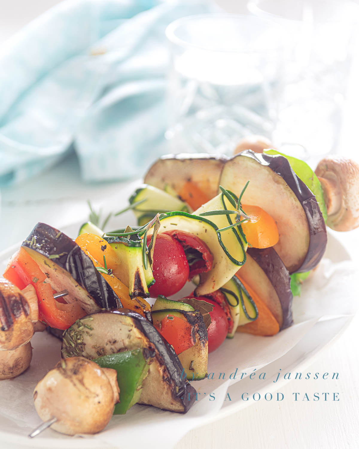 three vegan vegetable kebabs grilled, on a white plate. A Blue towel in the back