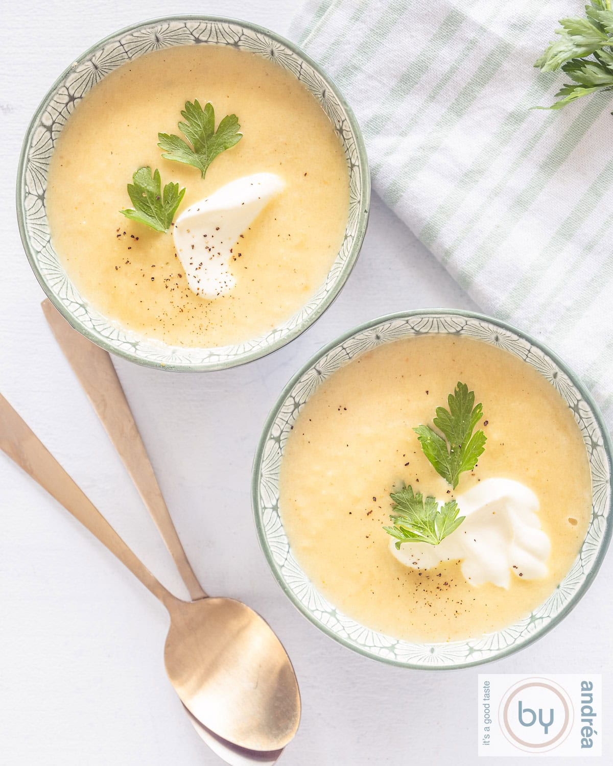Een foto van bovenaf van twee kommen gevuld met pastinaaksoep met yoghurt en koriander. Twee gouden lepels liggen eronder.