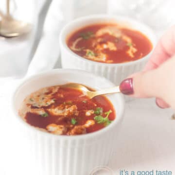 Two white bowls filled with tomato soup with meatballs and a dollop of cream. A hand with a spoon taking a bite.