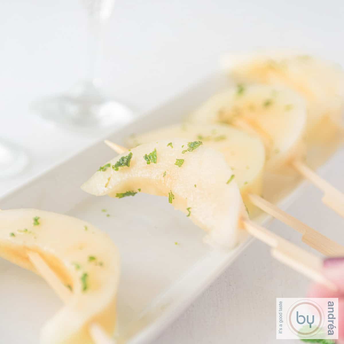 A square photo of a long white bowl of melon on a stick, marinated in a mojito cocktail. A hand grasps a piece of melon.