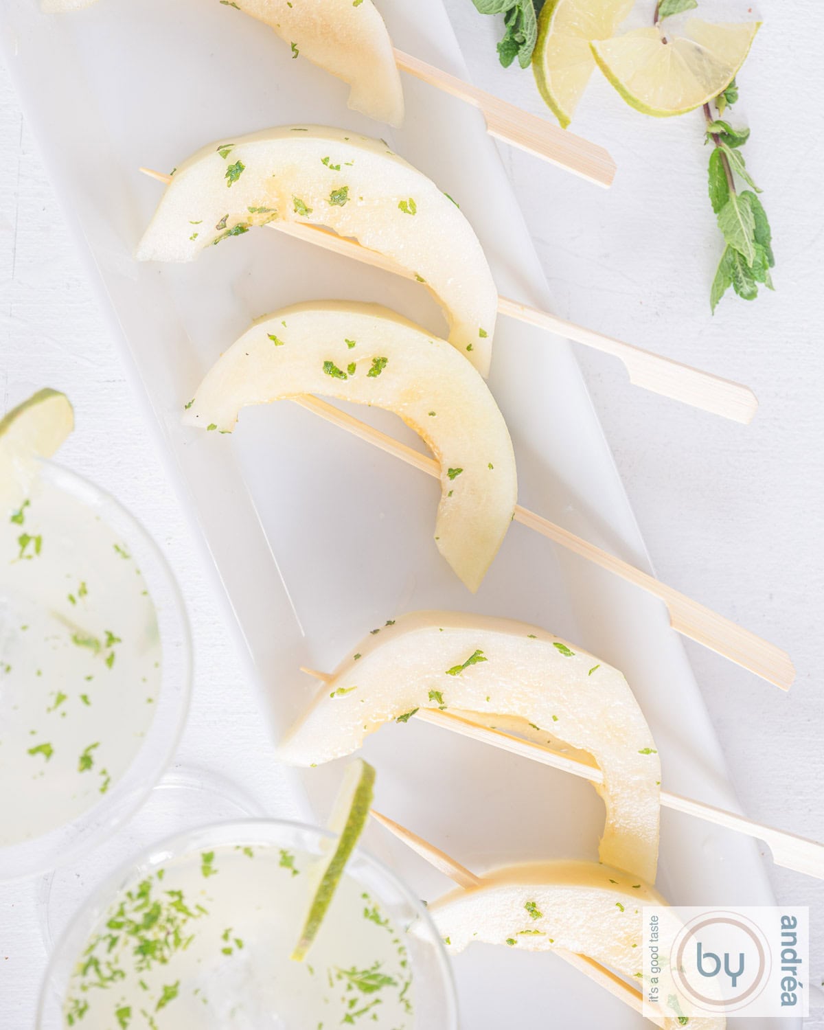 A top-down photo of a white bowl with mojito melon lollipops in a row. Two cocktails at the bottom. Some garnish at the top.