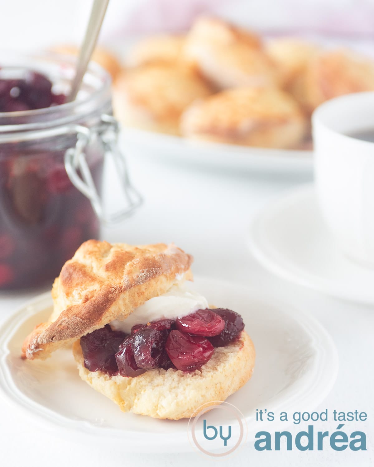 a sliced scone on a white plate topped with cranberry compote and clotted cream on a white background. In the back a jar of cranberry compote, scones and a cup of tea
