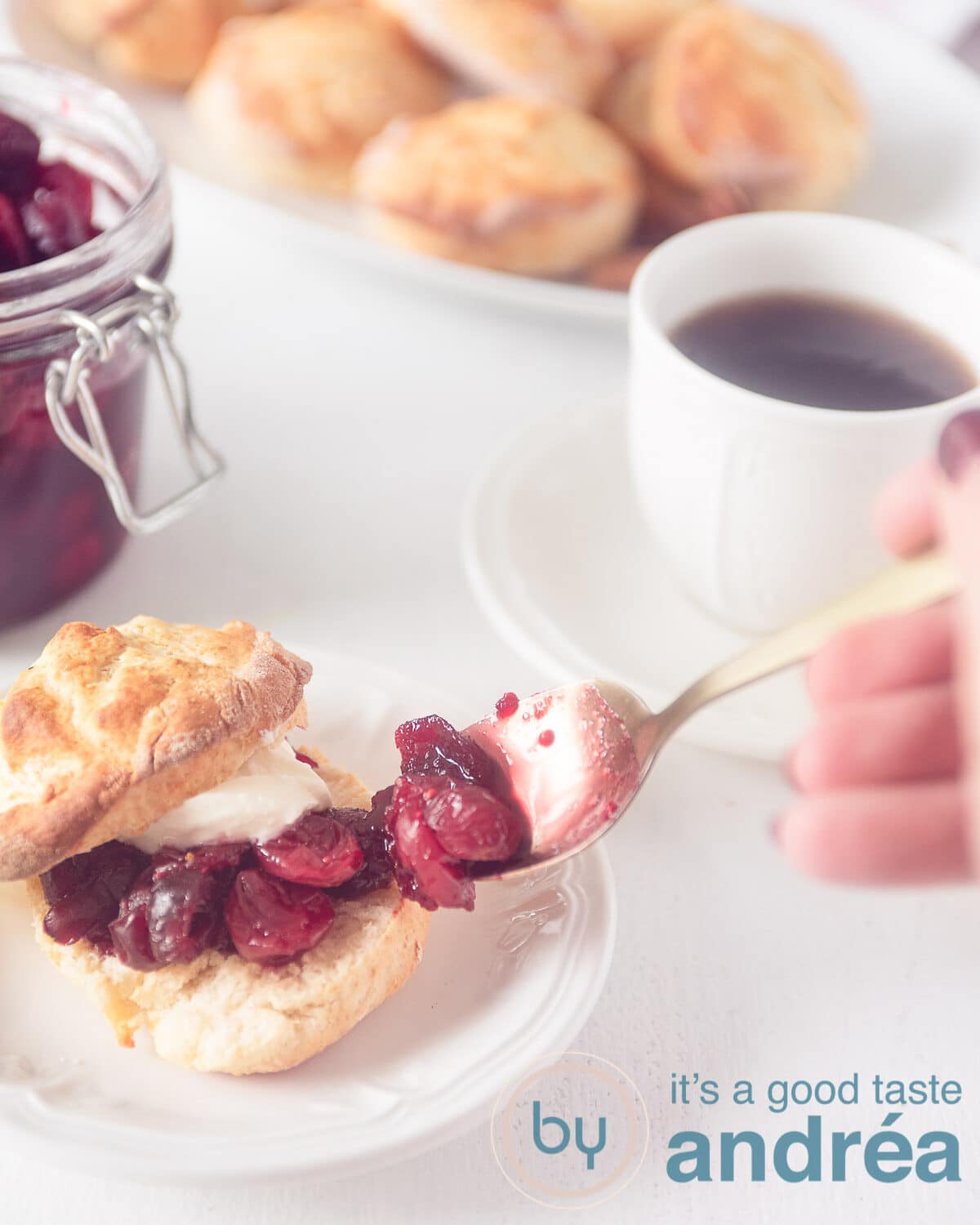 Een hand die extra cranberry compote op een opengesneden scone. In de achtergrond scones, een glazen pot met cranberry jam en een kopje koffie