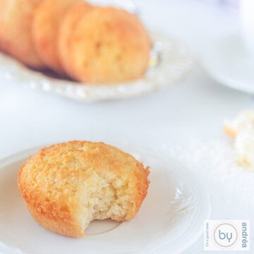 A square photo of a white plate with a coconut cupcake with a bite out of it. In the background a silver bowl with five extra cupcakes.