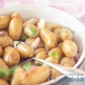 A square photo with a black and white oven dish filled with potatoes with skin, shallots and herbs. A spoon sticks into the bowl.