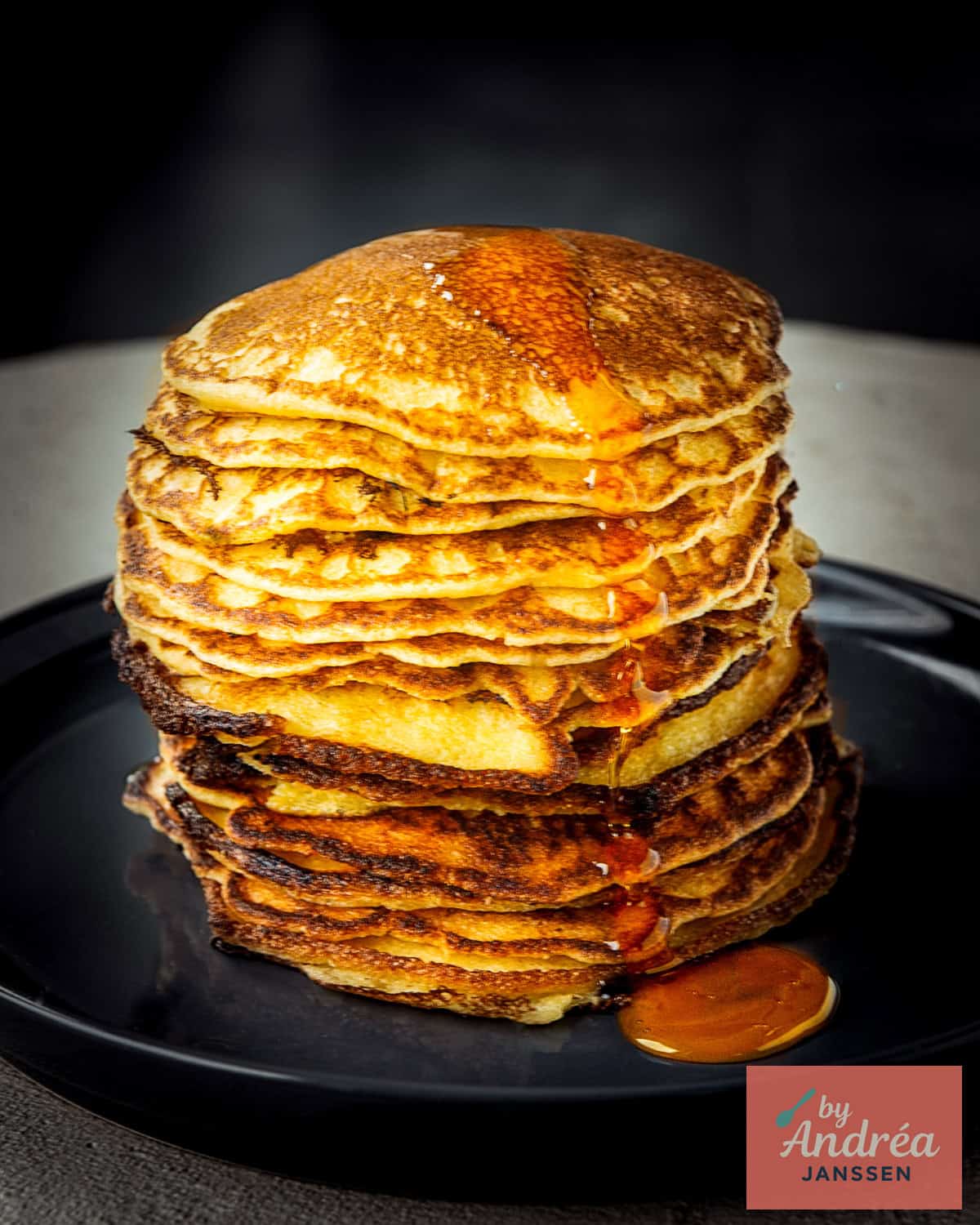 A black background a pile of buttermilk cornmeal pancakes and some maple syrup