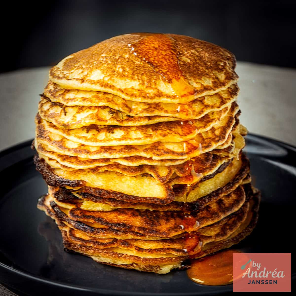 A square picture with a black plate a pile of buttermilk cornflour pancakes and some maple syrup drizzled on top.
