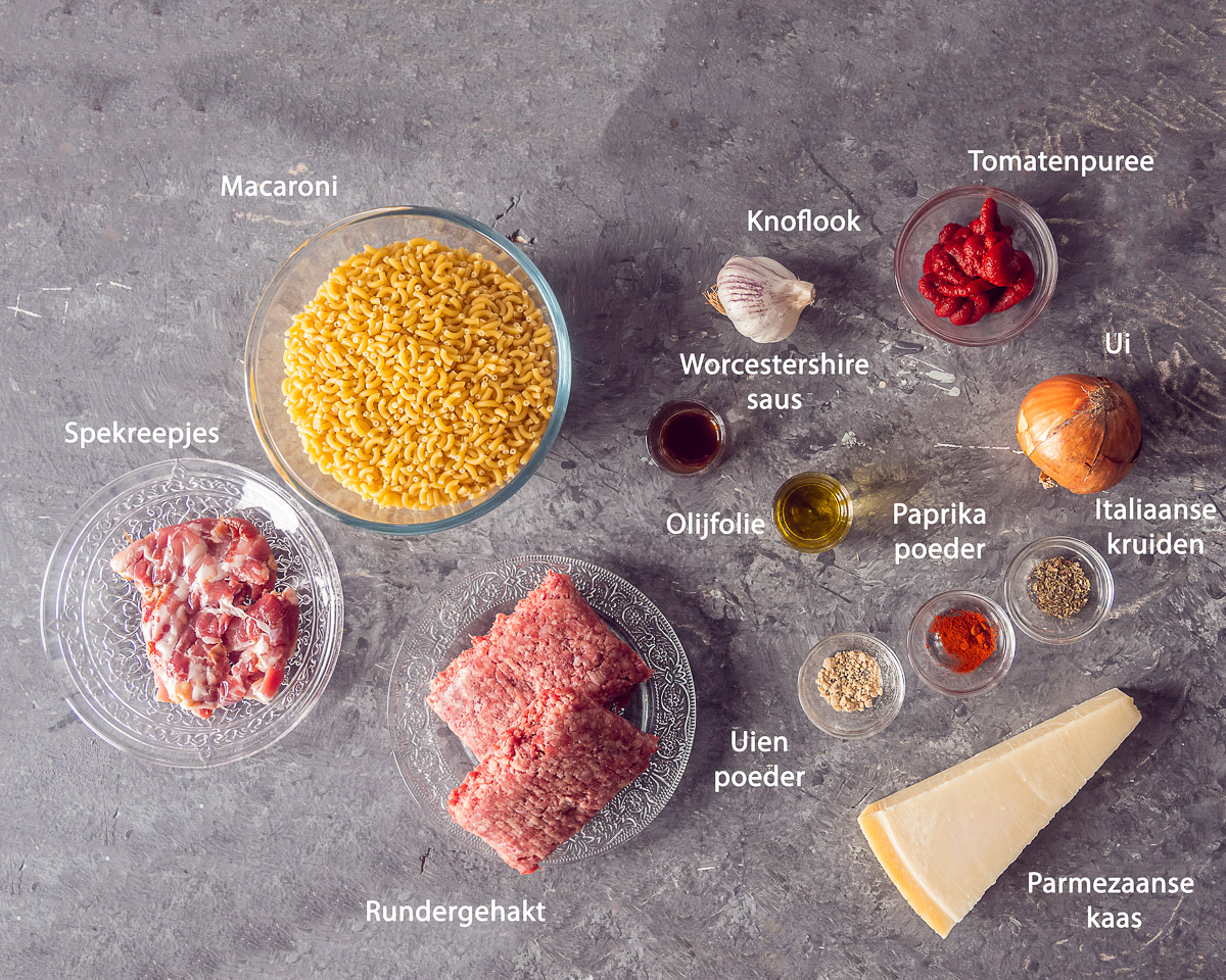 Ingrediënten voor macaroni met gehakt en spekjes op een grijze ondergrond, met Nederlandse namen bij de producten.