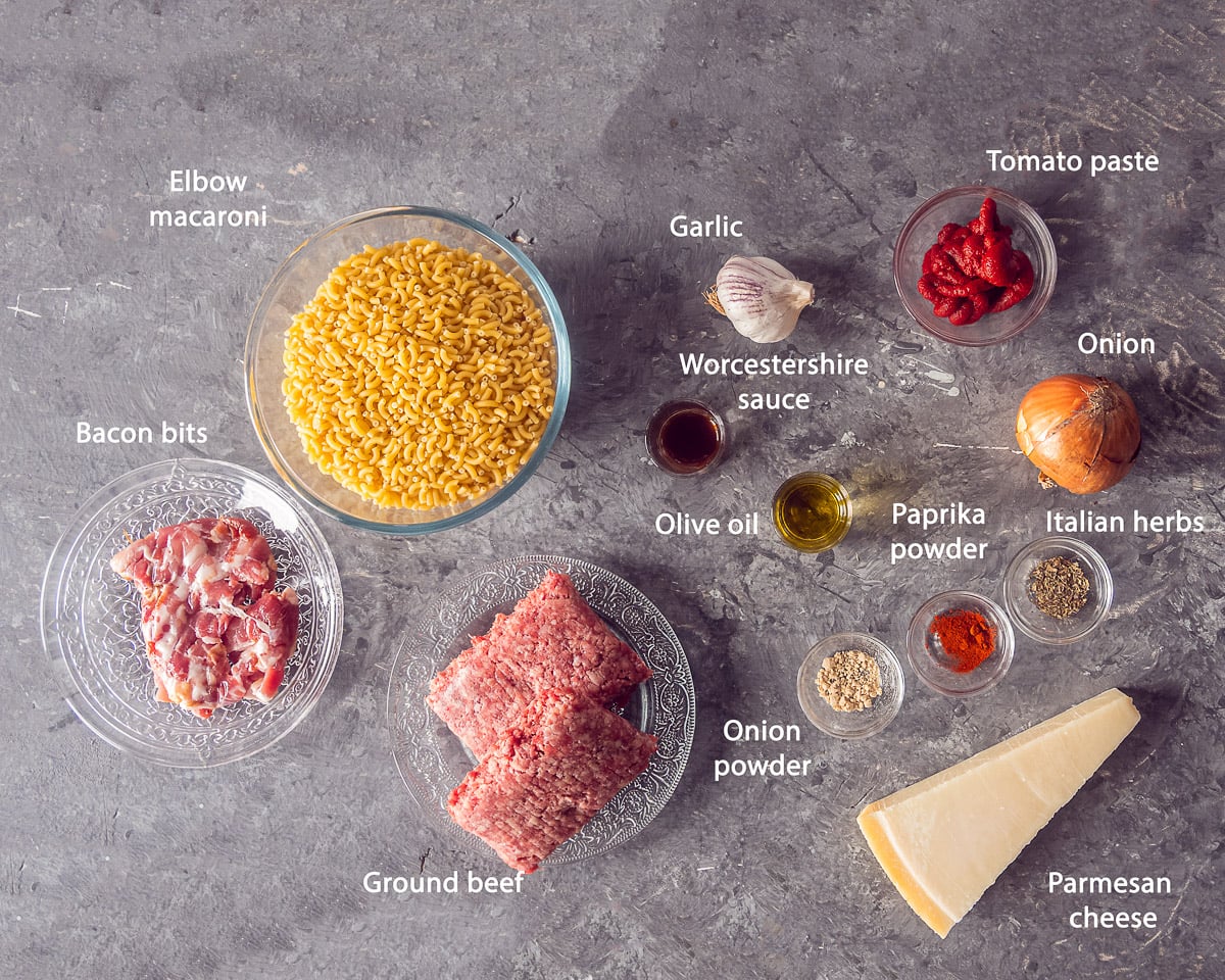 Ingredients for Dutch macaroni with ground beef and bacon arranged on a gray surface, labeled in English.