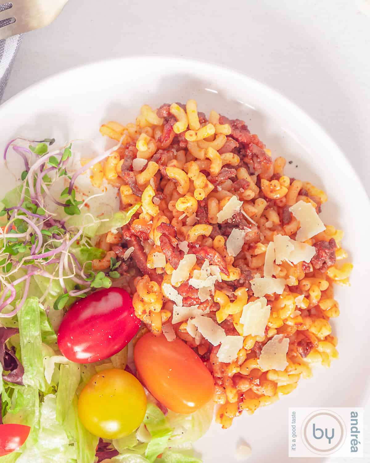 A top-down photo of a white plate with super-fast macaroni with minced meat, bacon and tomato on the right and a salad on the left