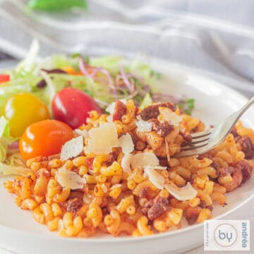 A square photo of a white plate with macaroni filled with onion, ground meat, bacon, and tomato paste. A salad is served on the side.