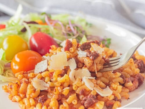 A square photo of a white plate with macaroni filled with onion, ground meat, bacon, and tomato paste. A salad is served on the side.