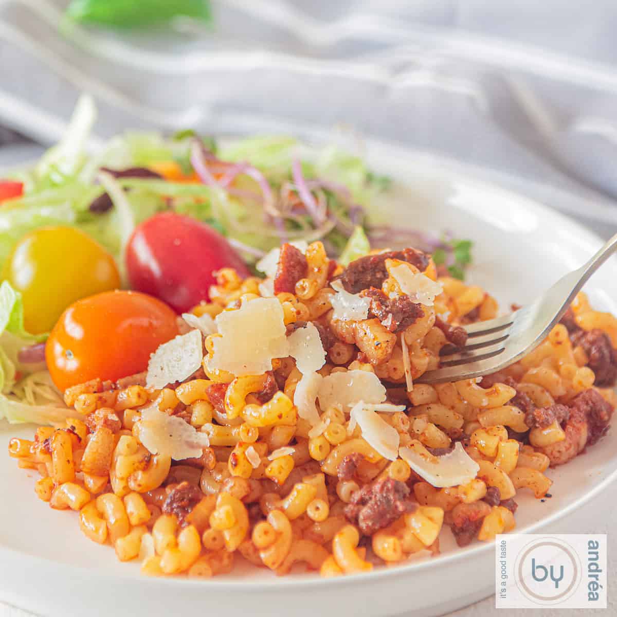A square photo of a white plate with macaroni filled with onion, ground meat, bacon, and tomato paste. A salad is served on the side.