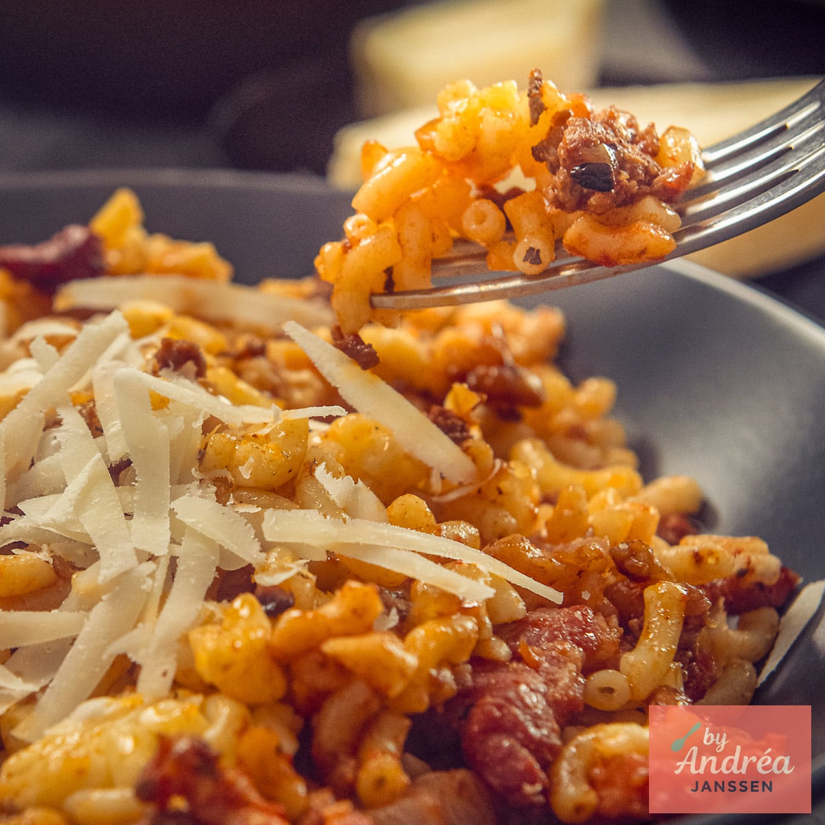 A forkful of macaroni and ground beef above a plate of macaroni with bacon and shaved Parmesan.