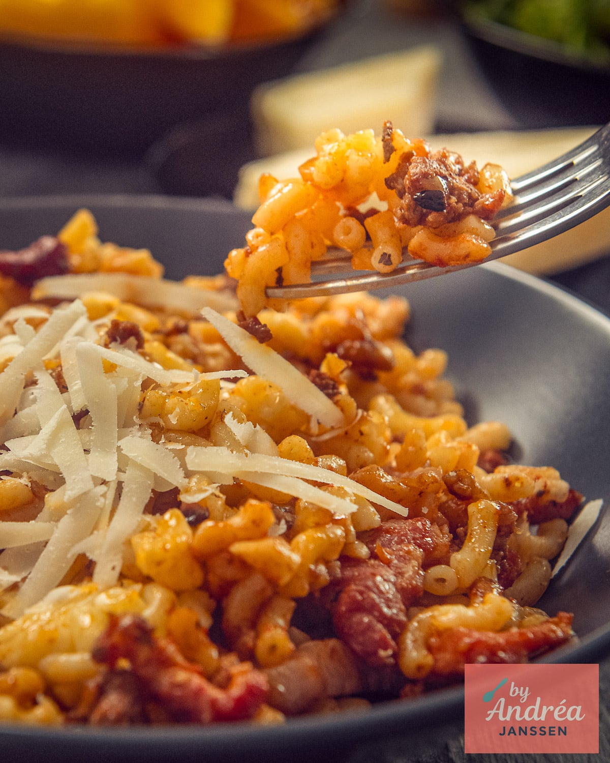 A forkful of macaroni and ground beef above a plate of macaroni with bacon and shaved Parmesan.