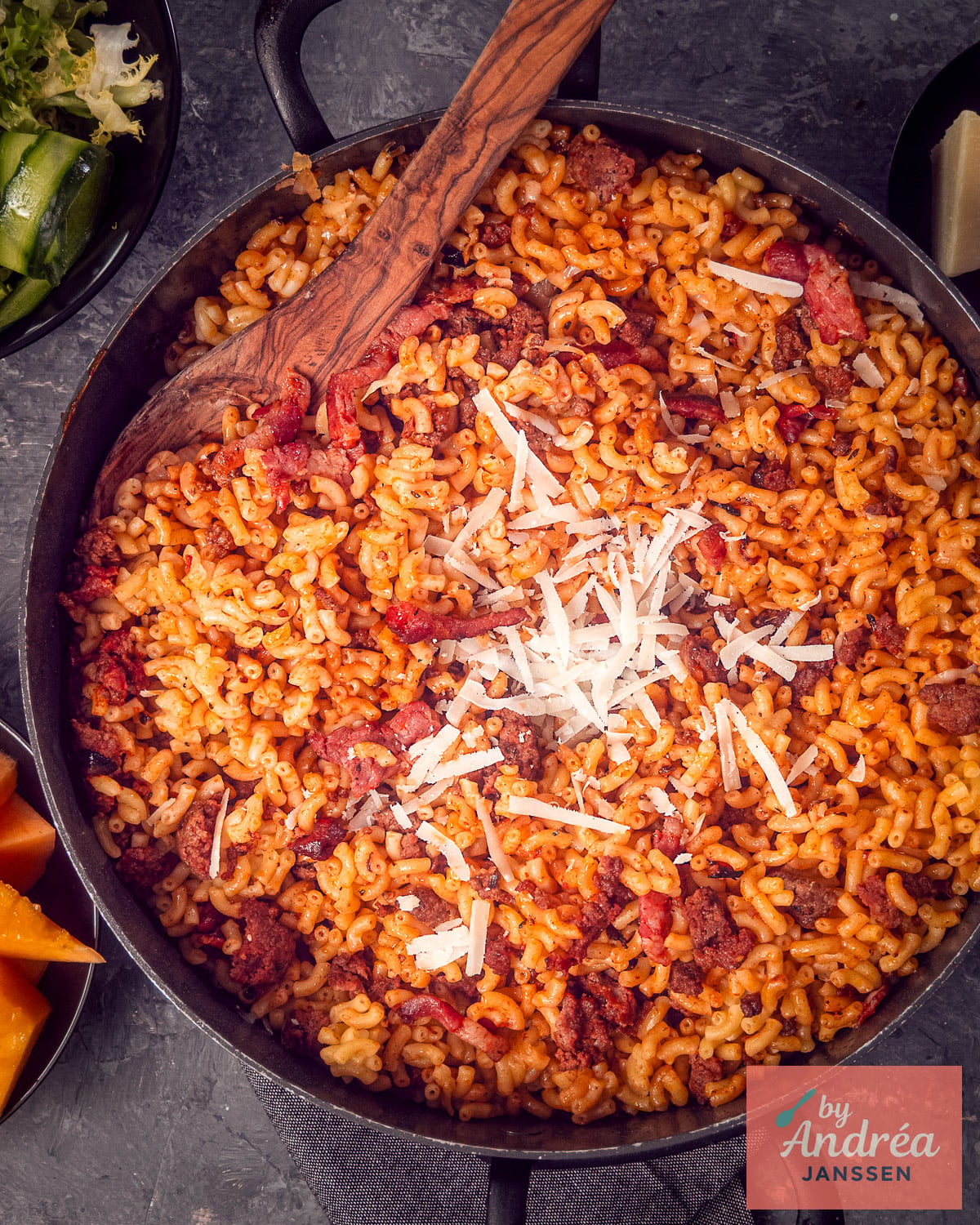 Overhead view of a pan of macaroni with ground beef, bacon, and Parmesan, with a wooden spoon resting inside.
