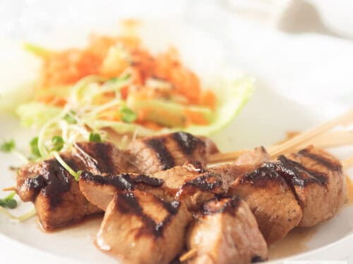 A square photo of a white plate with three skewers of pork tenderloin with ketjap and a salad with carrot and cucumber in the background