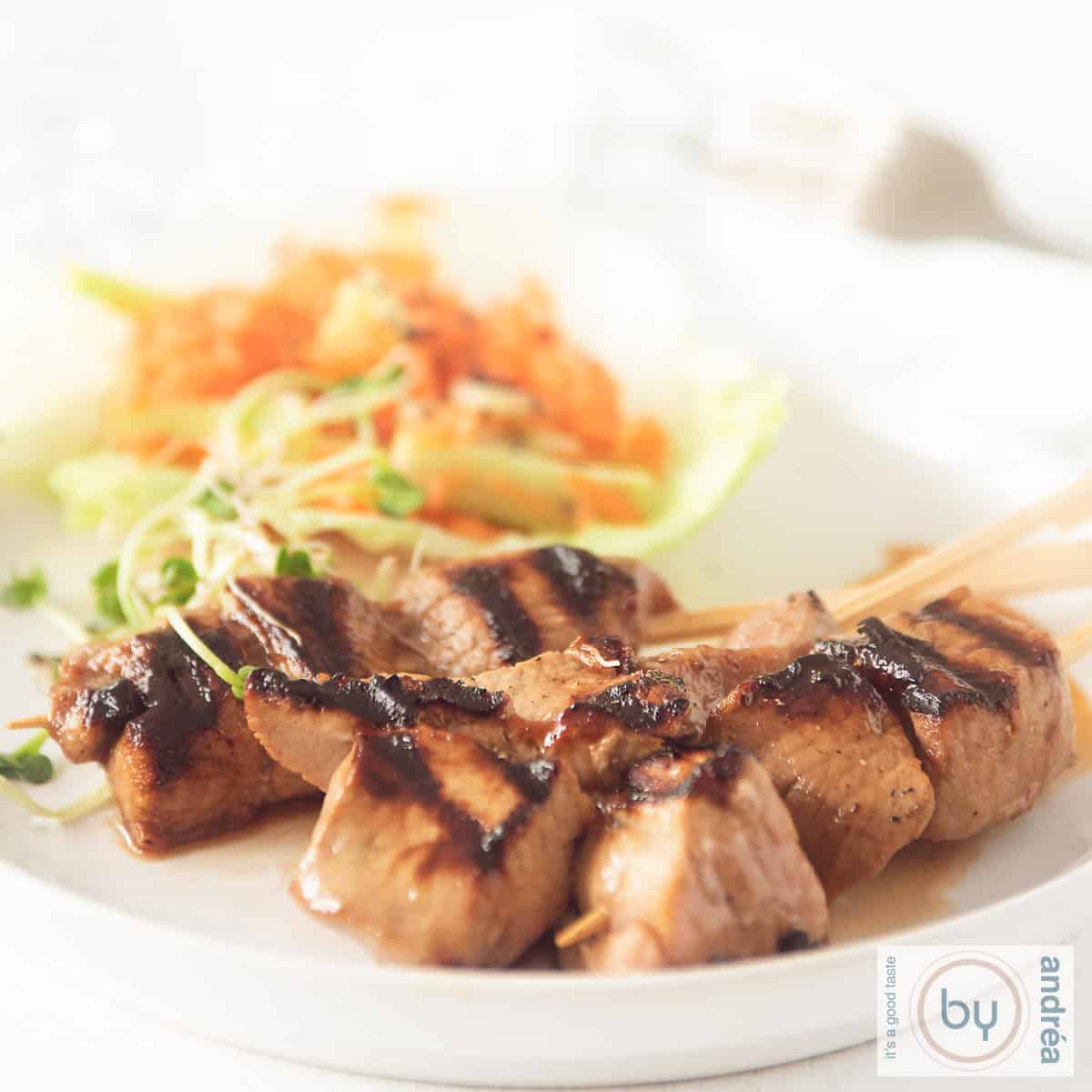 A square photo of a white plate with three skewers of pork tenderloin with ketjap and a salad with carrot and cucumber in the background