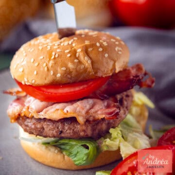 A square photo with a burger filled with feta on a bun with tomato, bacon, and lettuce.