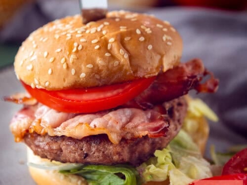 A square photo with a burger filled with feta on a bun with tomato, bacon, and lettuce.