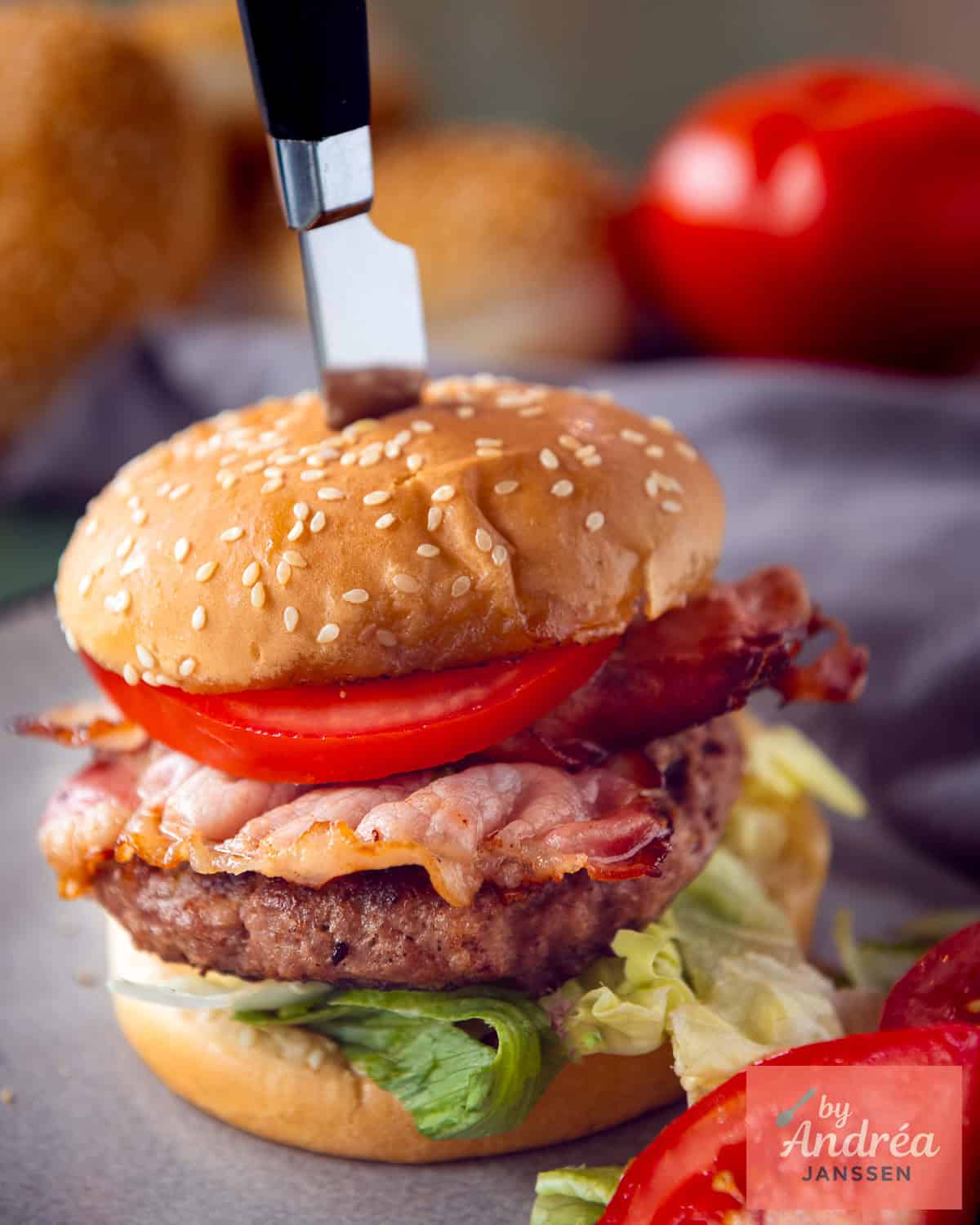 Een hamburger met feta op een broodje met bacon, sla en tomaat. Een mes steekt in het broodje