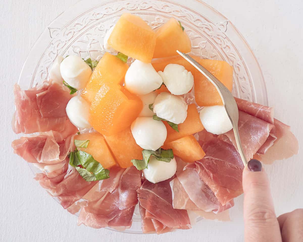 A glass plate with parmaham, mozzarella, melon, basil and dressing. A fork mixing everything.
