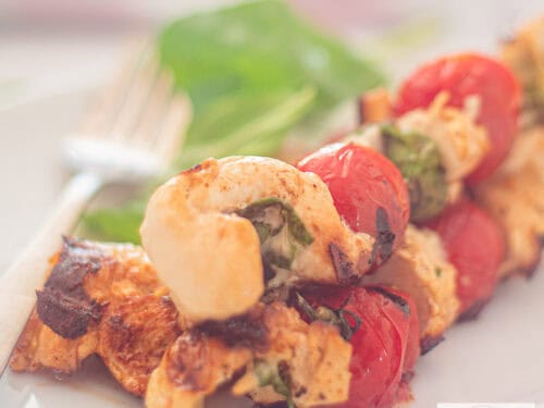 A square photo of three skewers with chicken, basil, mozzarella and tomatoes.