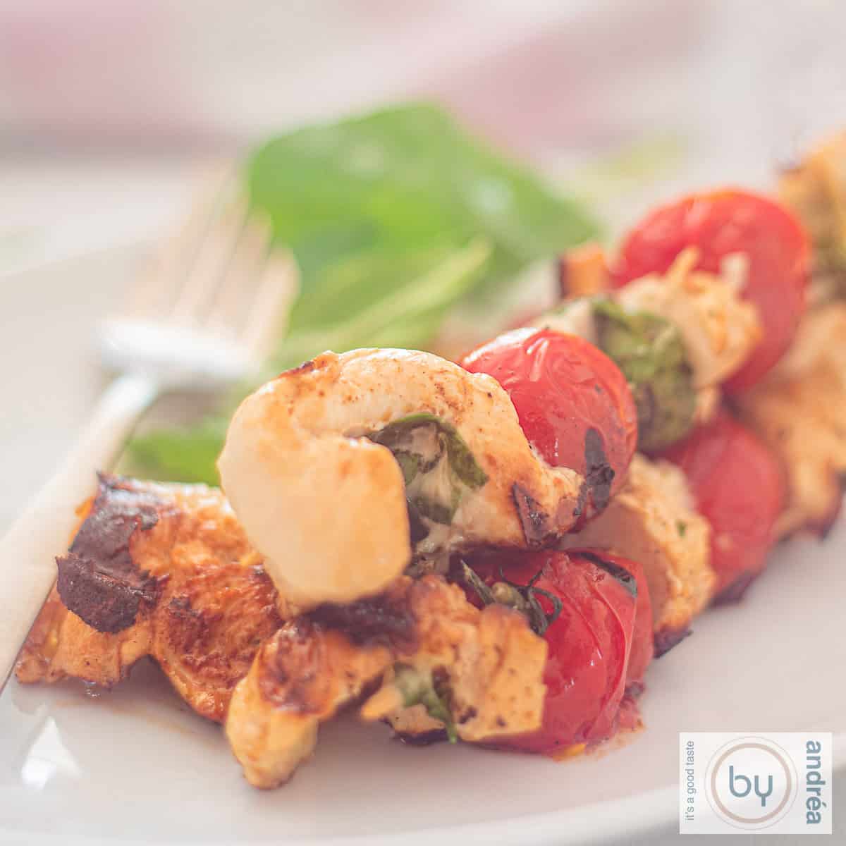 A square photo of three skewers with chicken, basil, mozzarella and tomatoes.