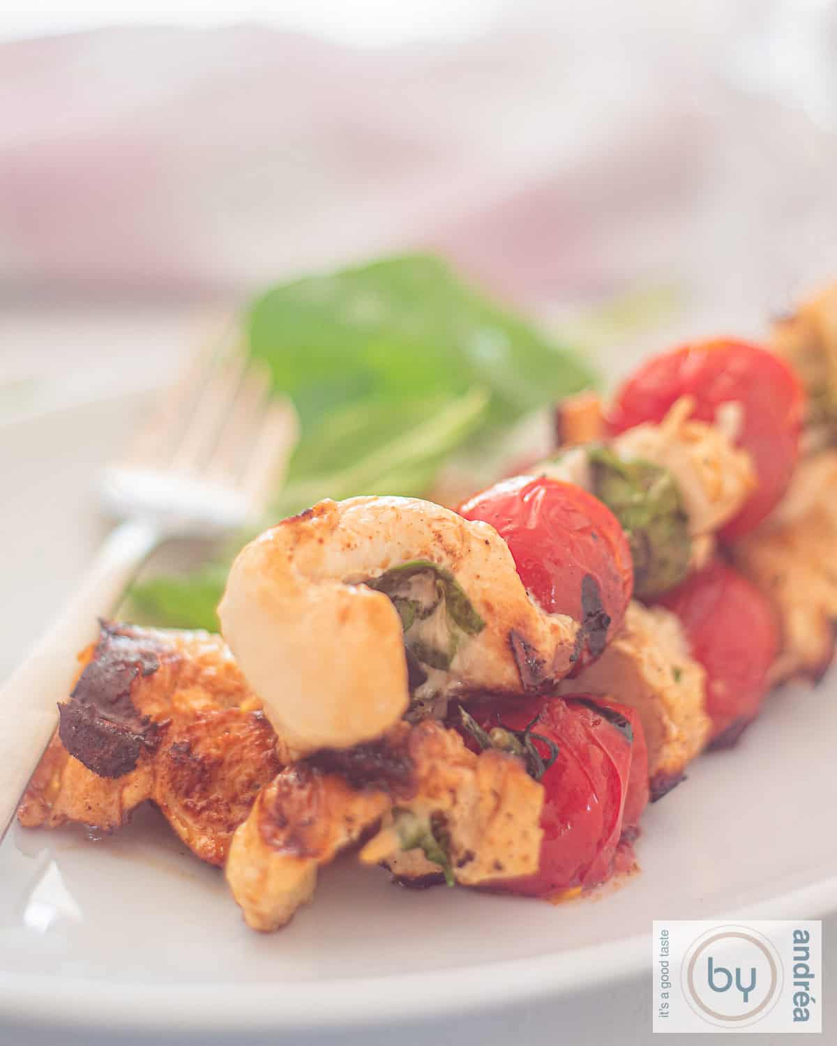A photo of a white plate with three skewers of stuffed chicken with mozzarella and basil and grilled cherry tomatoes.