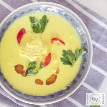 A square photo with a gray bowl filled with chicken soup with coconut milk, chicken and herbs.