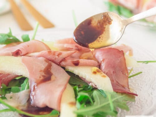 A square photo of a plate of rocket, pancetta and slices of melon. Dressing is drizzled on top!