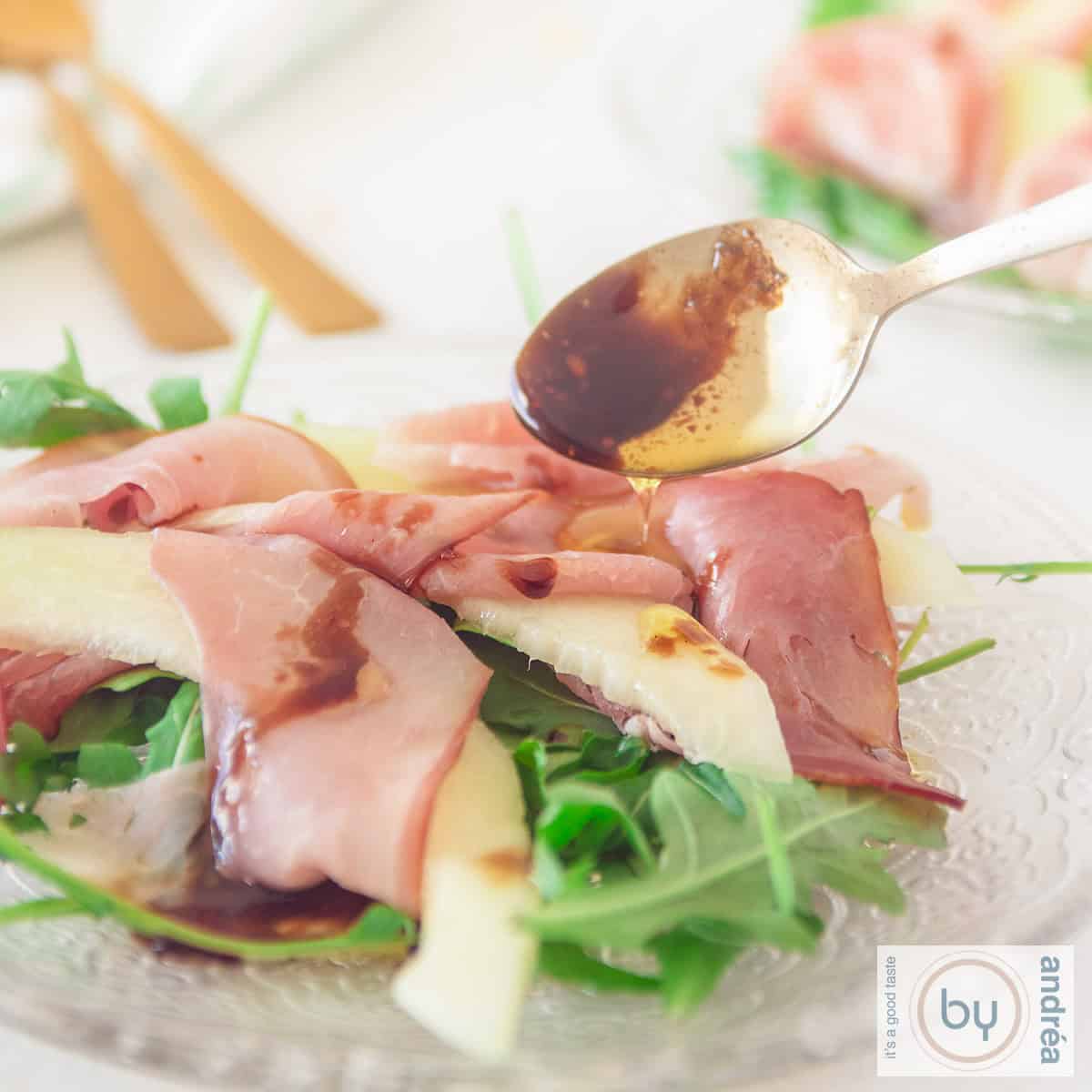 A square photo of a plate of rocket, pancetta and slices of melon. Dressing is drizzled on top!