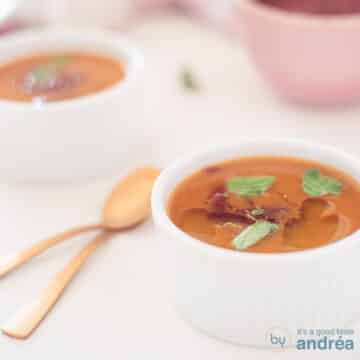 A square photo with a bowl of spicy pumpkin soup. A second one comes diagonally behind it. Two golden spoons to the left.