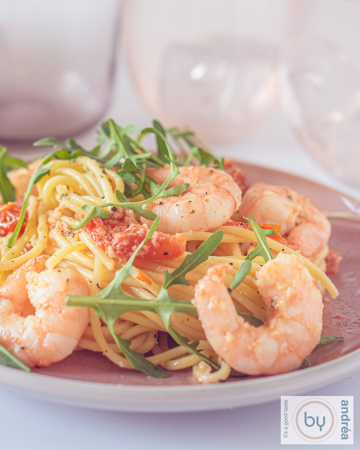 Een foto met een roze bord met spaghetti, rucola, garnalen en tomaatjes.