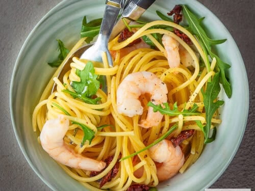 A square photo with a green bowl with pasta, shrimp and arugula