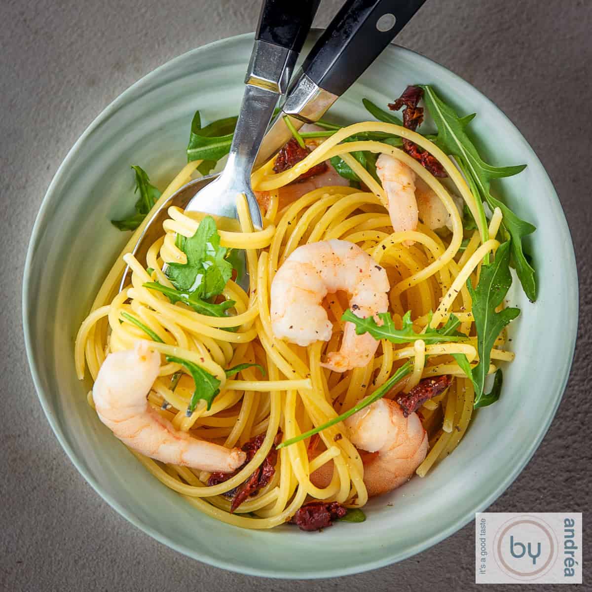 A square photo with a green bowl with pasta, shrimp and arugula
