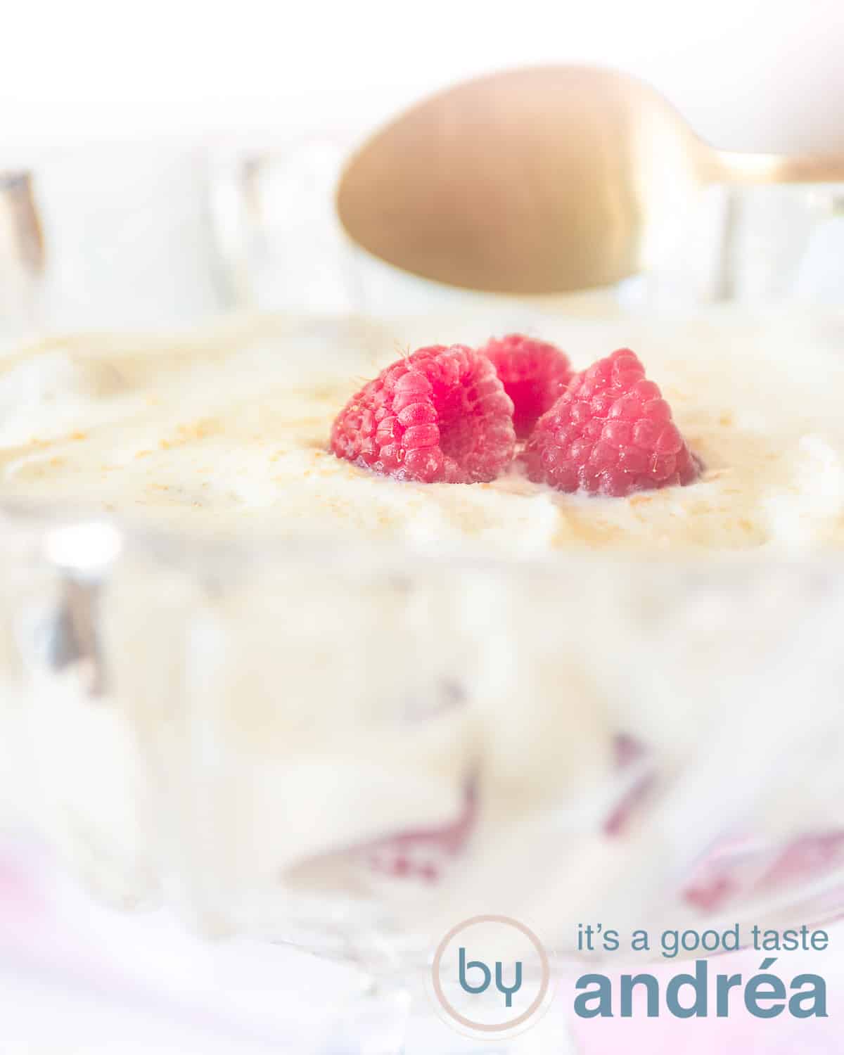 A glass bowl filled with raspberries, lady fingers and whipped cream. Topped with fresh raspberries. A golden spoon wants to take a bite