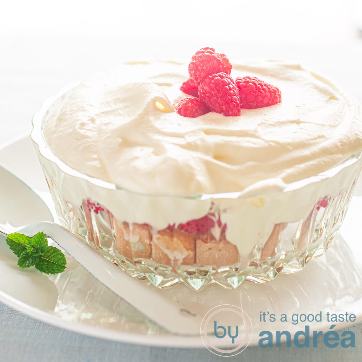 square photo with a glass bowl with lady fingers, raspberries and whipped cream. Topped with fresh raspberries