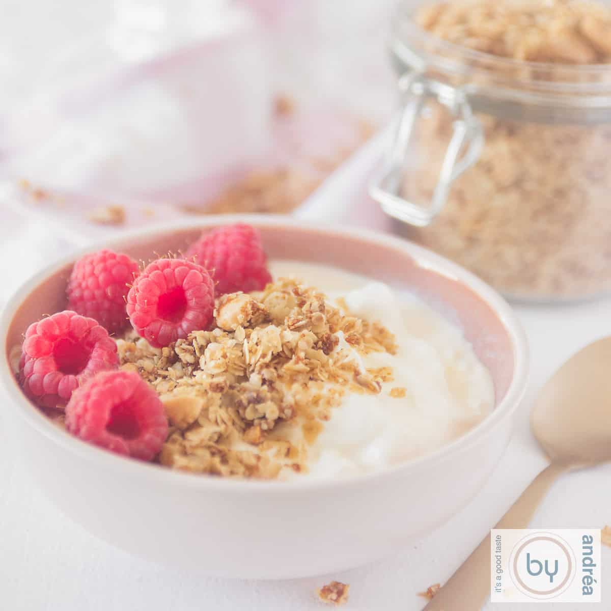 A square picture with a pink bowl filled with yogurt, raspberries and almond granola. Some more granola in the background.