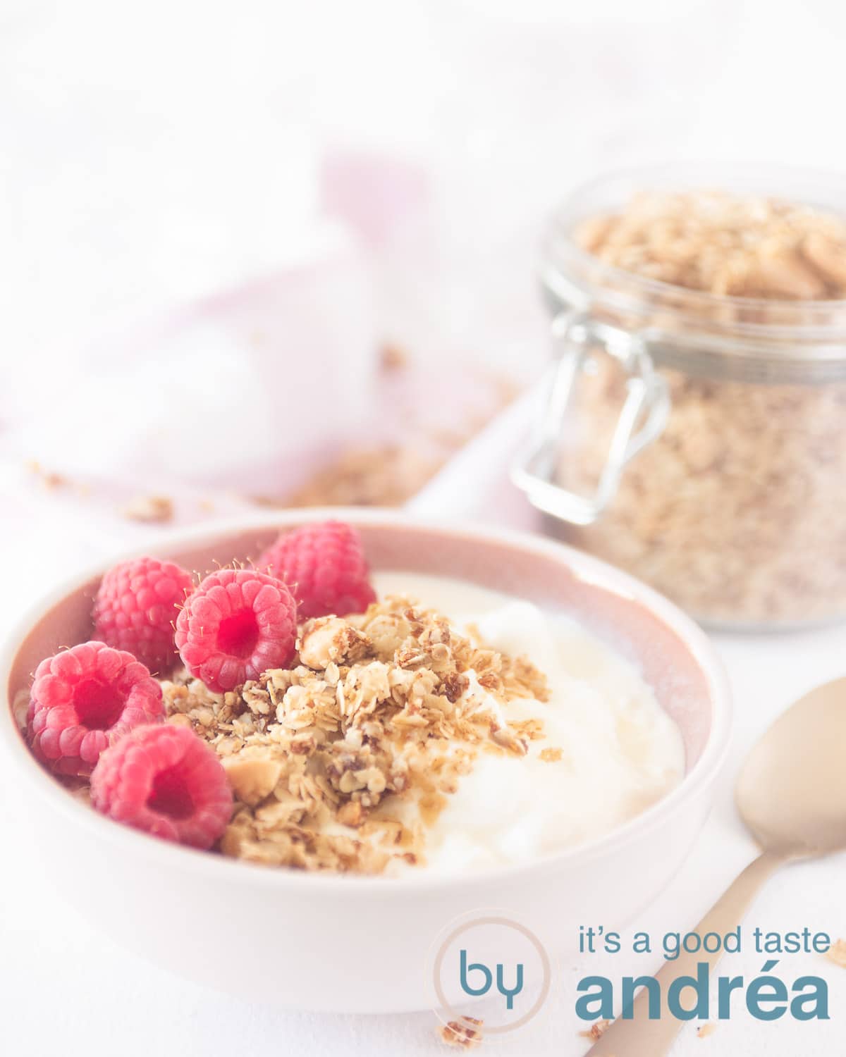 a pink bowl filled with yogurt, granola and raspberries. In the right back a jar with granola. a golden spoon on the right.