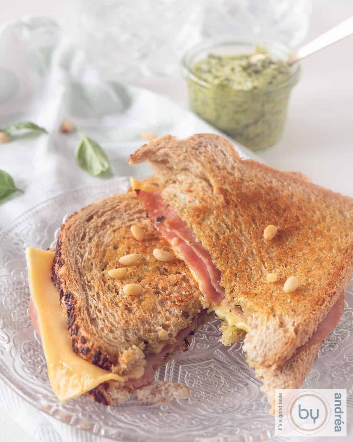 Een glazen bord met een tosti in tweeën gesneden, met pijnboompitten. In de achtergrond een potje pesto.