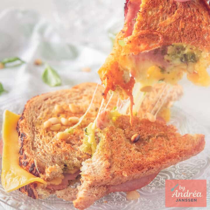 A square picture with two slices of a sandwich with cheese, pesto and ham on top of each other, cheesy strings visible