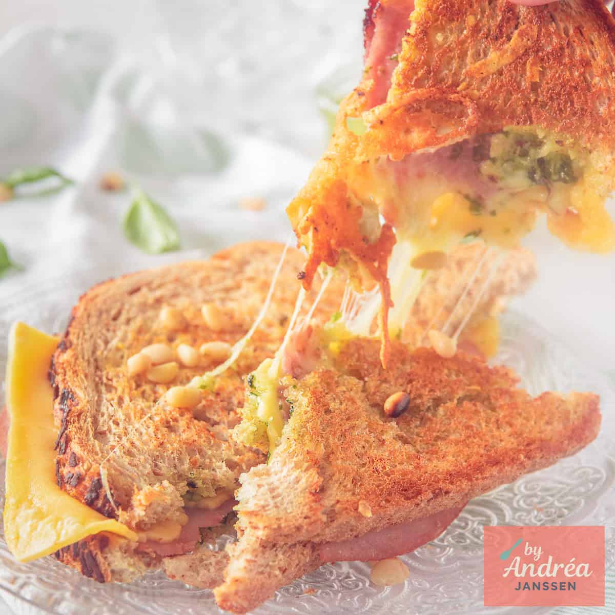 A square picture with two slices of a sandwich with cheese, pesto and ham on top of each other, cheesy strings visible