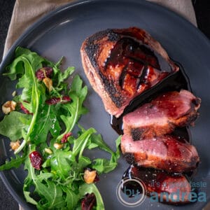 A square photo of a gray plate with duck breast fillet, 3 slices cut off and a balsamic sauce with honey