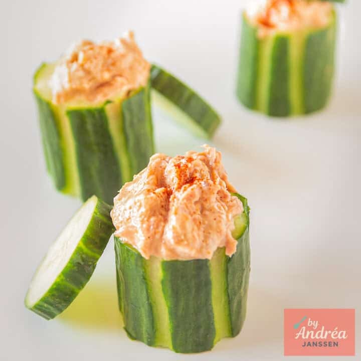 A square photo with three cucumbers filled with tuna salad, with a cucumber cap against it.