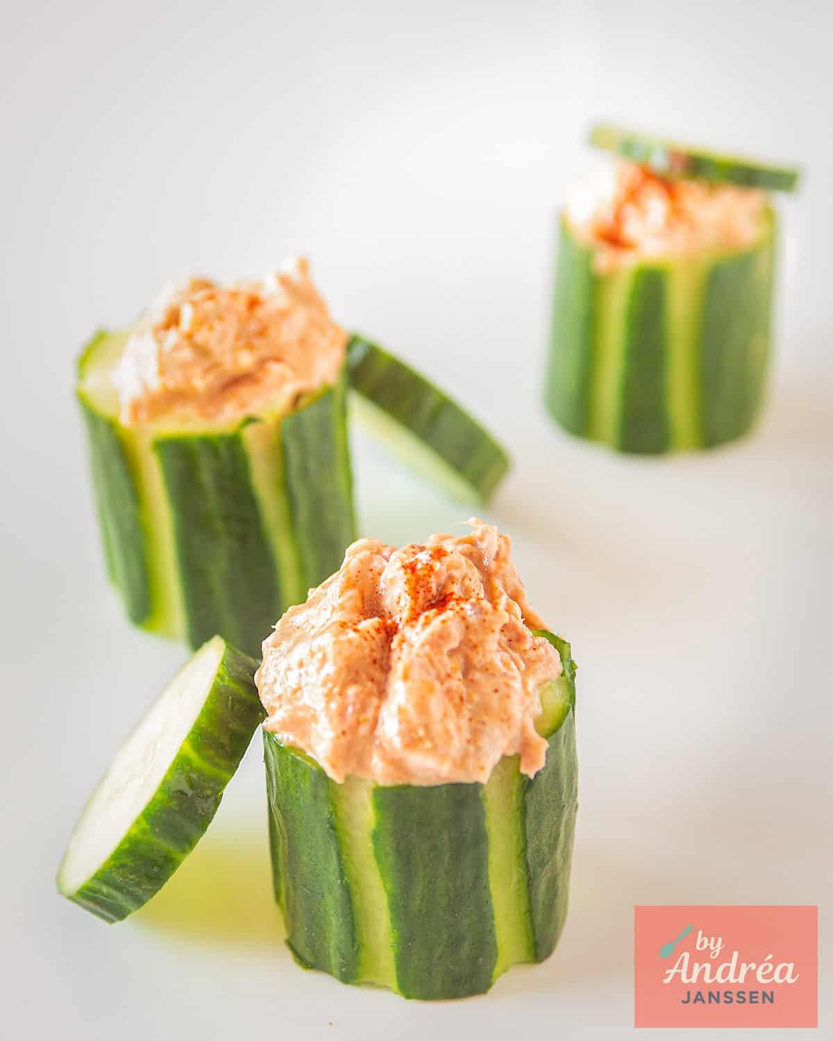 Three cucumber bites in a row on a white plate with tuna salad and a cucumber cap.