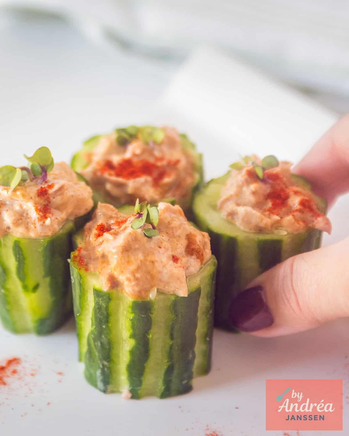 Four cucumbers filled with tuna salad and paprika. A hand takes a cucumber bite.